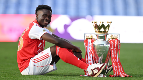 Arsenal's Bukayo Saka sitting next to the Premier League trophy