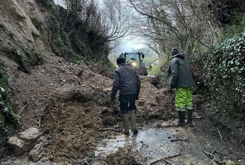 Two men stand in a small road in Somerset looking at a huge pile of earth and rocks that has blocked the road