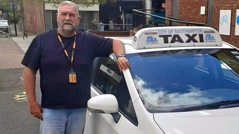 Perry McMillan wearing a t-shirt and jeans and leaning against his parked taxi which says "city of Southampton taxi" on the top of it.