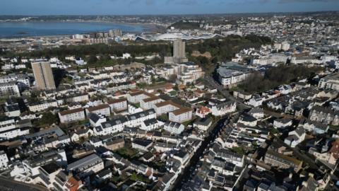 An aerial shot of houses in South St Helier in Jersey in the Channel Islands. A mixture of houses and larger blocks of flats with a number of roads in between. The sea is also in the background. 