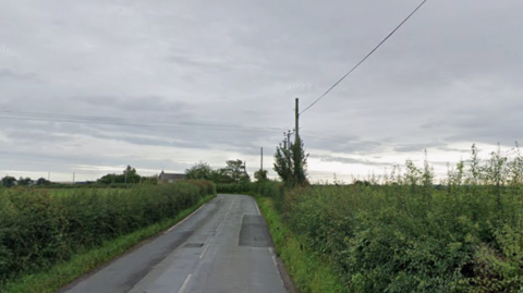 A Google Maps picture of the B3092, a country lane with hedges on either side and a power line on the right side of it.