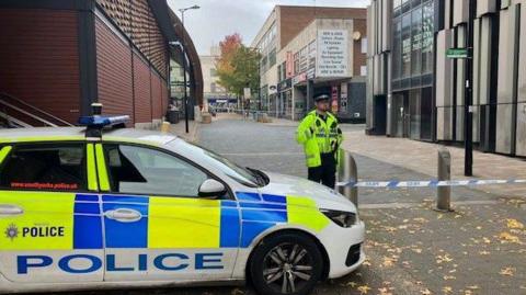 A police officer stands by a cordon marked with police tape at the end of a road, next to a police car.