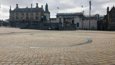 Queen Victoria Square in Hull city centre. Spiralling brick work on the vast space of floor in the foreground with three historical buildings with steps leading up to them in the background.