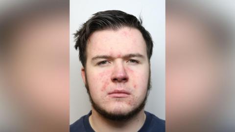 A mug shot of a man with brown hair, a short brown beard and spots on his face. He is wearing a navy t-shirt and is frowning.