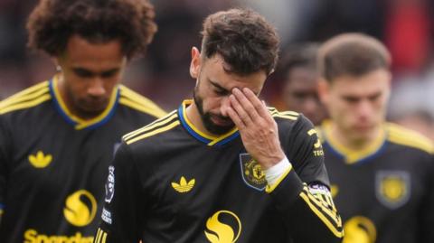 Manchester United captain Bruno Fernandes with his left hand on his head on the pitch after losing to Brentford. There are other players behind him as they walk over to thank the travelling Manchester United fans