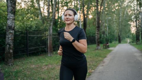 A woman wearing headphones and a dark grey t-shirt jogging on a path in a park with trees around her