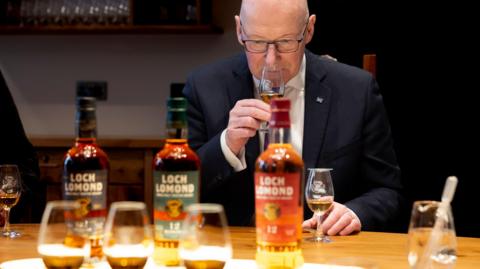 John Swinney is a bald man wearing glasses and a dark suit. He holds up a glass of whisky to his nose and appears to sniff it. A couple of drams and bottles of Loch Lomond whisky sit on a table in front of him.