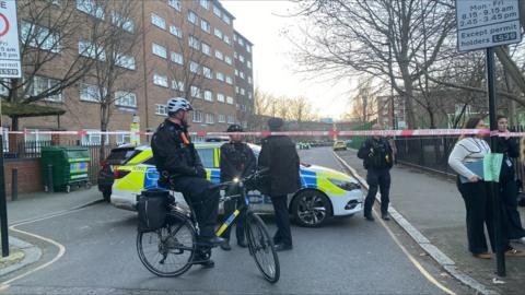 Police car and officer on bike beside red police cordon with further police activity down the road