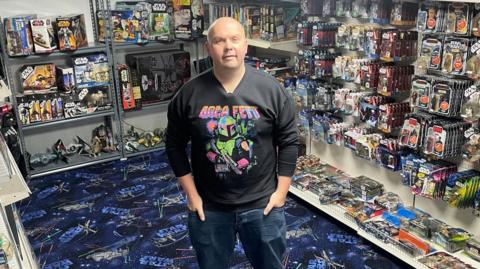 A bald man in jeans and a sweatshirt stands among hundreds of sci-fi figures and spaceships neatly displayed in rows.