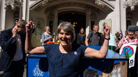A woman wearing a dark blue shirt is standing outside a Supreme Court. There are campaigners holding banners standing behind her.