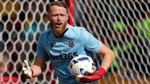 Stoke City goalkeeper Viktor Johansson in his blue shirt holding the ball in his left hand as he prepares to restart the play