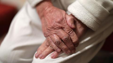An elderly person's hands rest in their lap. They are wearing white trousers. Their hands are free from any rings or nail paint.