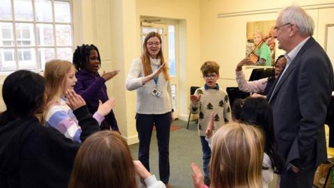 A group of young people standing around in a circle singing. They are in a yellow music room. 