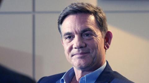 John Michie wearing a dark blue suit jacket and blue shirt. He has short dark hair.