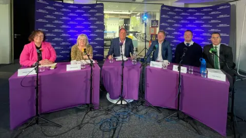 The five candidates sitting in a row behind tables with presenter Chris Mann in the middle.