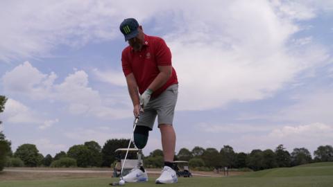A man with a prosthetic leg stood with holding a golf club about to hit a golf ball.