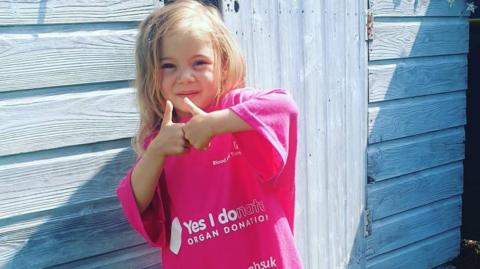 Five-year-old Thalia-Beau Wright stands in front of a blue slatted wooden shed with her thumbs up. She has long blonde hair and wears an oversized pink T-shirt from an organ donation charity, with white writing and logos across the front.