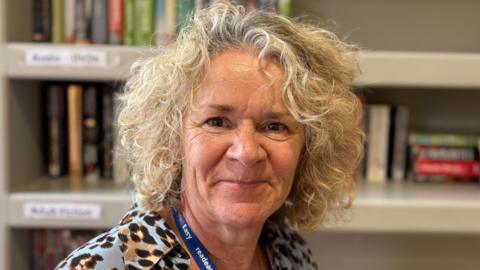 Rachael Eddy with long curly blond hair smiling at the camera and wearing a black, brown and blue top. There are books on shelves behind her.