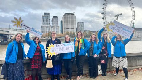 Eight women wearing blue jackets and holding signs reading "Lingen Davies Sunflower Appeal" stand in front of the London eye and high-rise buildings with the River Thames behind them.