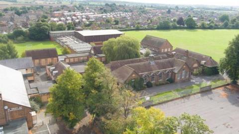 A school site pictured from above. An assortment of buildings can be seen, as can a playground and large playing field. In the far distance, rows of houses are visible.