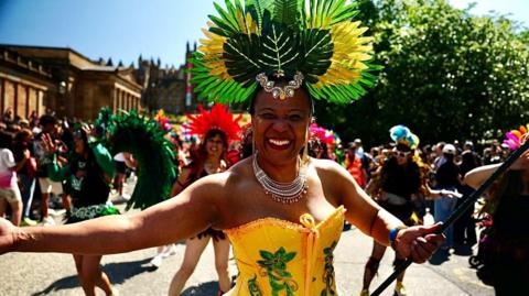 A carnival performer wearing a large green and yellow feathered headdress and a yellow-and-green costume walks at the front of a colourful street parade.