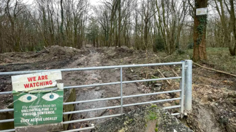 A silver gate on a path into Hoads Wood with a sign on saying: 
WE ARE WATCHING
Fly tippers face an unlimited fine or a prison sentence.
CCTV ACTIVE LOCALLY