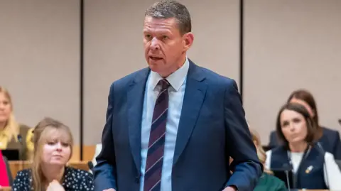 Rhun ap Iorwerth stood in the Senedd chamber surrounded by his colleagues, who are sat down. He is speaking.