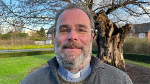 Rev Cawthorne looking into the camera, he's wearing a grey jumper and you can see he's wearing a vicar's clerical collar underneath. Behind him you can see some of the affected homes on Abbot Drive, and to his right is an oak tree.