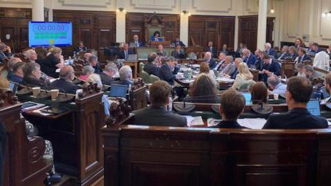 A packed hall of local councillors attending a meeting, sit in a circle facing inwards on wooden seating.  An electronic countdown clock is on the wall. 