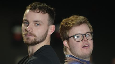 Callum Foster with dark short hair and Edward Wagland with blonde short hair stand back to back while looking at the camera. The man with dark hair is also wearing a black T-shirt while the other man is wearing a blue top with black glasses.