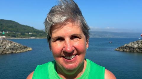 Elaine is smiling into the camera. The sea is behind her. She's wearing a green top.