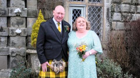 Kenny Macloed (left) standing alongside Lisa. He is bald and wearing a black jacket and yellow kilt. Lisa has blonde hair and is wearing a light blue dress. She is holding a small bouquet of flowers in her left hand. They are in front of an old stone building with leaded windows.