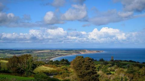 An ariel shot of the north of the Isle of Man, you can see the arching coastline, and there are houses scattered on the land amongst a lot of fields.