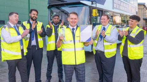 Six bus drivers wearing yellow high-vis jackets, one of whom is holding a certificate