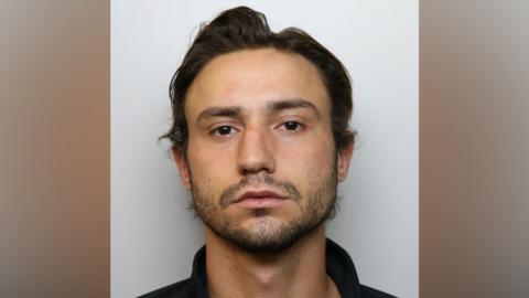 A man with dark hair and a beard appears in a police mugshot. He is wearing a black polo shirt.