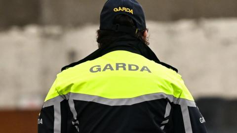 A garda officer photographed from behind.  They are wearing a navy cap and a navy jacket with fluorescent shoulders and reflective silver stripes
