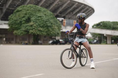 A young female BMXer sits on top of her bike in a car park outside of a stadium in Lagos