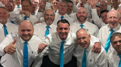 A number of men are pictured together smiling and wearing white shirts and blue ties. They are arm-in-arm.