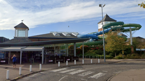 The entrance to Leisure World. It is a modern looking building with a mostly glass facade. However, off to the right is a tower from which a green and a blue flume come out of and lead down towards the back of the building.