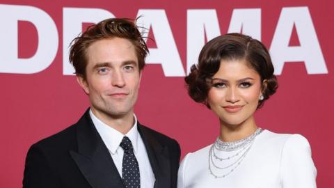 Robert Pattinson and Zendaya smiling at the camera. He has brown hair and is wearing a black suit jacket, black tie with white polka dots and a white shirt. Zendaya is wearing a white dress and diamond earrings and necklaces. She has short brown hair.
