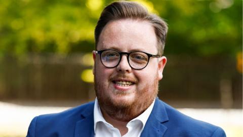 Thomas Kerr, a man with dark glasses, light brown hair and a ginger beard, smiles in a close-up shot. He is wearing a blue suit and white shirt, with blurred-out vegetation in the background 