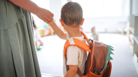 A child is holding their parent's hand as they go into a room with other children. They are wearing a backpack.
