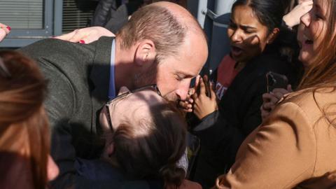 Prince William hugs a shocked Natasha Gorry who holds her hands up to her face and laughs. Prince William, who is considerably taller appears to be talking to her as he smiles. Ms Gorry is wearing black rimmed glasses and her light brown hair is tied back. She is standing at the front of a crowd of people who are all smiling.