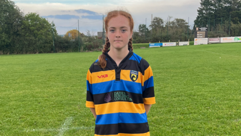 Mega Board standing on a rugby field wearing her yellow, blue and black striped rugby shirt. She has her hair in two braids and is looking at the camera with a serious expression.