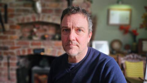 Scott Farr wears a navy jumper and is looking directly at the camera. The background is blurred but you can see a bare brick open fireplace behind him and a picture from up on a sage green wall. 