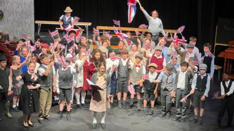 Schoolchildren are dressed in clothes from the 1940s as they sing on a stage with the light shining on them as they wave British and Jersey flags.