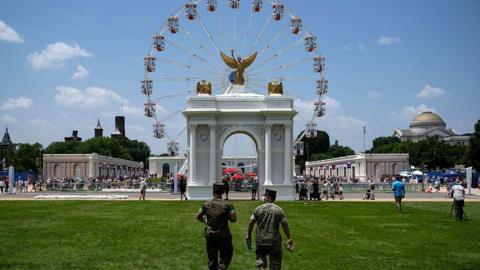 a view of two men walking through fair grounds in front of mock triumphal arch and ferris wheel