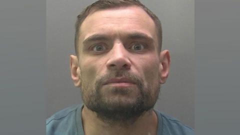 A police mugshot of Wayne Sharman. He has short light brown hair and  a cropped beard. He is wearing a grey T-shirt and looking at the camera.