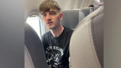 Tyler Kerry sits in a seat on a aircraft. he has short brown hair and a thin moustache. he is wearing a black T-shirt