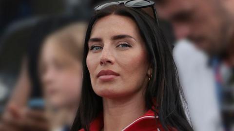 Annie Kilner, with long black hair, is wearing earrings. Black sunglasses are balanced on her head. She is wearing and a red-and-white jacket.